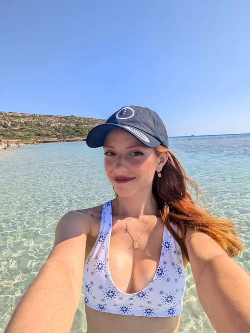A red-haired woman wearing a navy baseball cap and a white crocheted bikini top takes a selfie while standing in clear turquoise water at a beach.