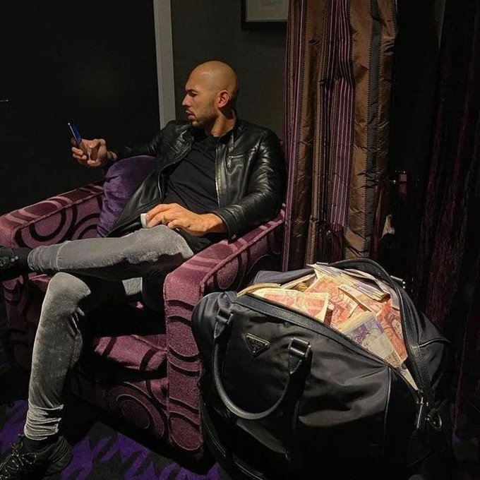 A bald man in a black leather jacket and gray jeans sits in a purple patterned chair, looking at his phone, while a black duffel bag overflowing with foreign currency sits next to him.