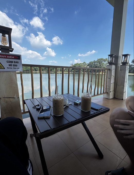 Two iced coffee drinks sit on a dark wooden table on a lakeside balcony, accompanied by phones and a small bag, with a 'CẢNH BÁO HỒ SÂU' warning sign nearby.