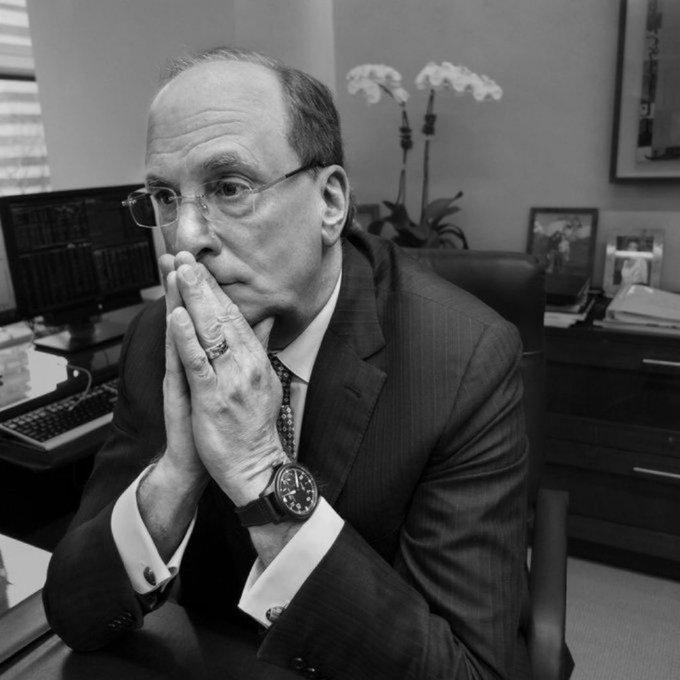 A black-and-white portrait of an older man in a suit and tie with glasses, sitting at a desk with his hands clasped together near his face in a thoughtful pose, wearing a wristwatch and ring, with orchids visible in the background.