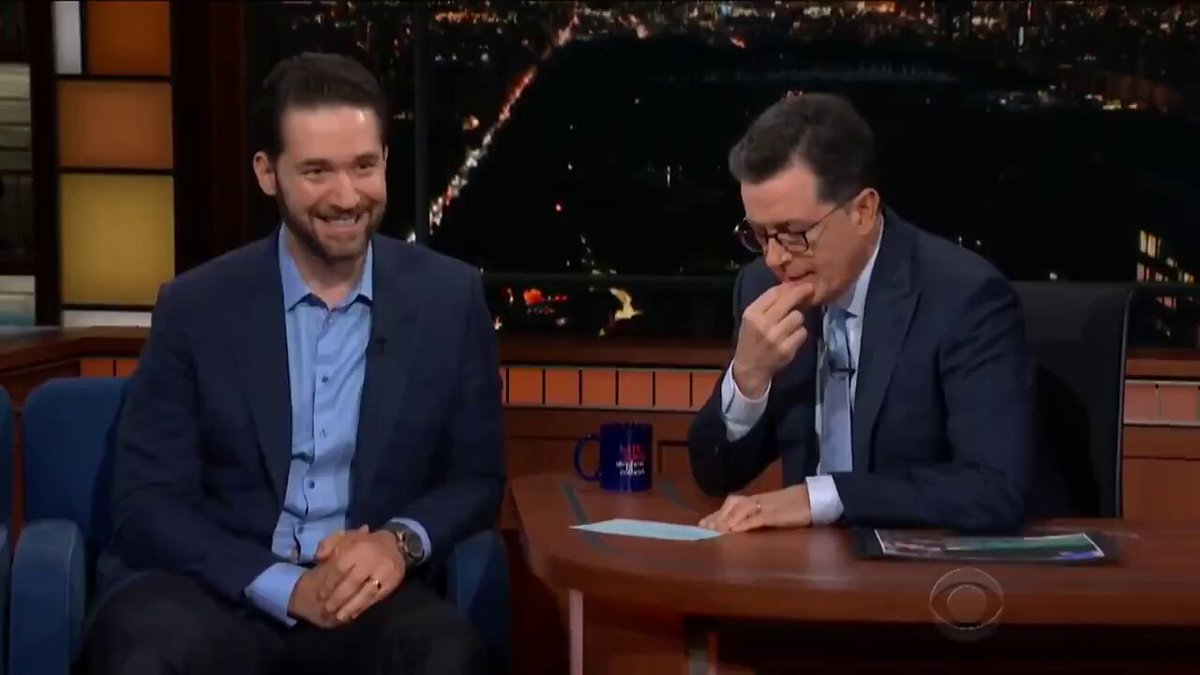 The image shows two men sitting at a desk, with one man wearing glasses and holding his nose while the other man has a smile on his face. The scene appears to be set during an interview or discussion about Bitcoin's importance in the digital age.