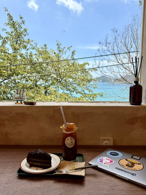 A chocolate cake slice and iced coffee sit on a tray by a window overlooking the ocean in Busan, with a laptop covered in stickers including one reading "XIII" placed nearby on the table.