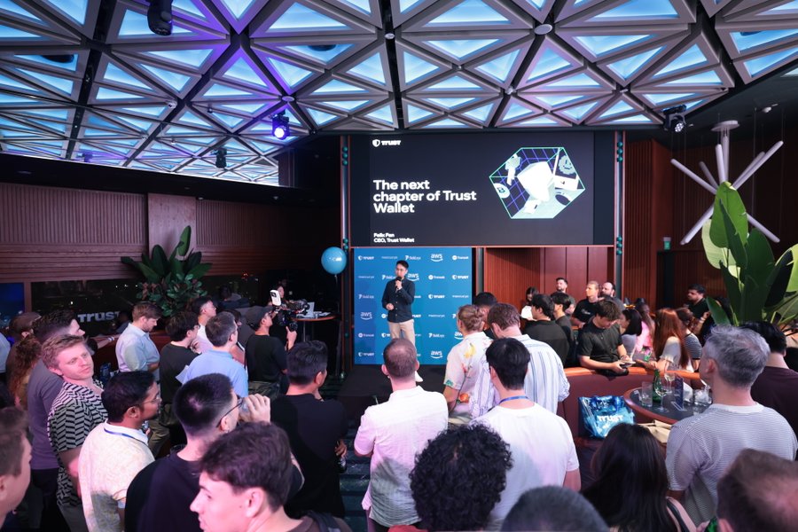 A presenter on stage at Token2049's "Beyond 9YA" event stands before a large screen displaying "The next chapter of Trust Wallet" while a packed audience of builders, founders, and industry attendees listens attentively. The image captures the full-house atmosphere referenced in the post, showcasing the dense crowd of Web3 innovators gathered under an illuminated geometric ceiling.