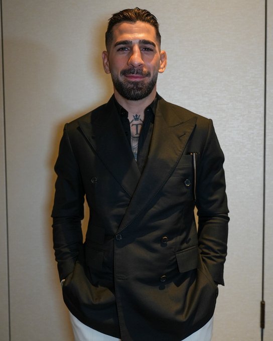 The image features a man wearing a black suit jacket with white pants, standing against a white wall. The man has a tattoo on his neck and appears to be posing for the camera. The caption of the image includes information about an event at the UFC White House on Saturday, June 14th at 8pm, featuring a fight presented by @CryptoCom.