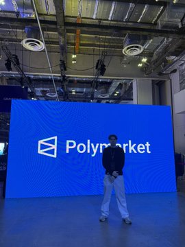 A person stands on a stage in front of a large blue screen displaying the Polymarket logo.