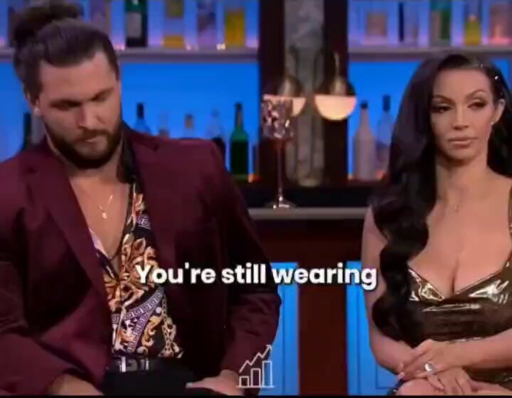 The image features two people, one man wearing a red jacket with a gold pattern and another woman dressed in a gold dress sitting at a bar. The text "You're still wearing" appears above them, which could be interpreted as a humorous or playful statement about their relationship status.