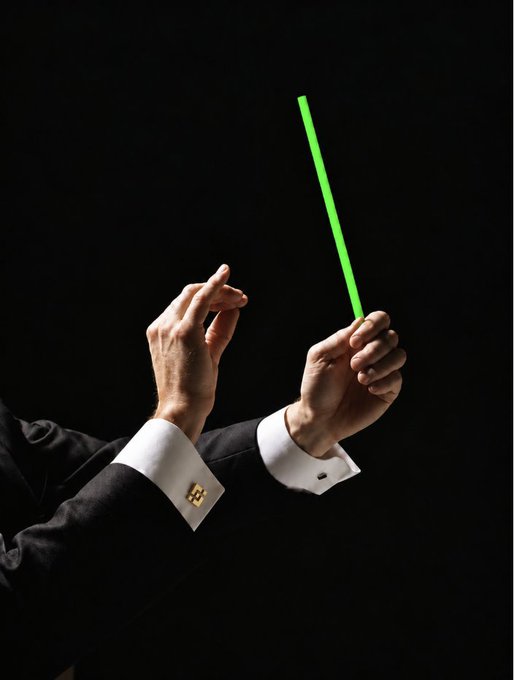 A man in a black suit with white French cuffs and gold cufflinks holds a bright green baton against a black background, poised to conduct. The image complements the post "Day 625，各就各位！继续演奏上涨的乐章！" by visually symbolizing the conductor's command to continue the bullish "music" of rising markets on Day 625.