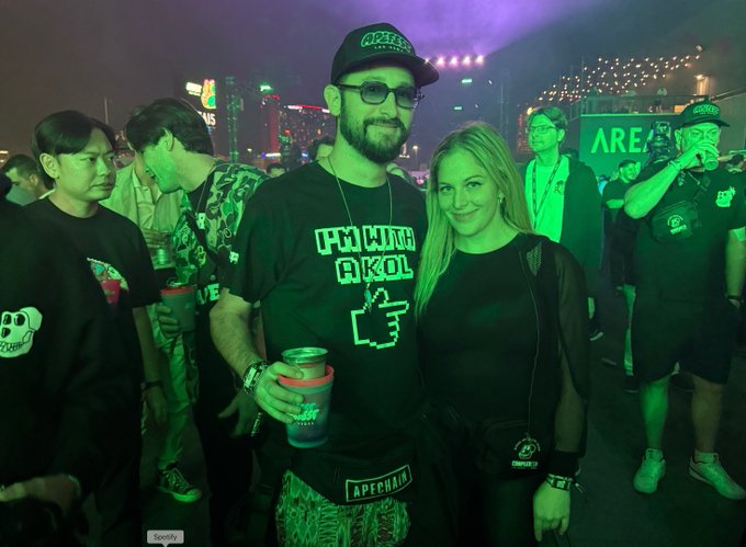 A nightlife photograph captures a bearded man wearing sunglasses and a cap alongside a blonde woman, both in dark attire, at what appears to be an ApePark event bathed in green lighting.