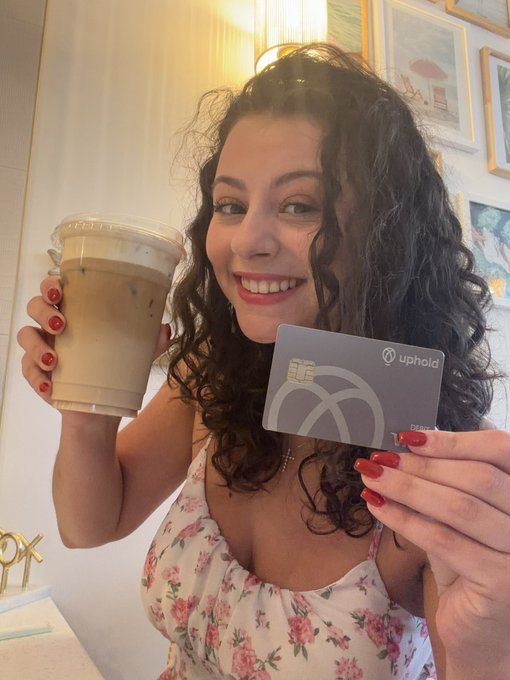 The image features a woman holding an open plastic cup with a beverage, possibly coffee or iced tea, along with a wallet containing two credit cards and some coins. She appears to be posing with her wallet for a photo. The post that she is promoting contains birthday wishes, specifically for someone's 14th birthday. XRP, a virtual currency, is being suggested as the form of payment for purchases made on this special occasion. The woman is emphasizing the benefits of using XRP by showcasing how it can be used to enjoy her favorite beverage while receiving rewards through her purchase.