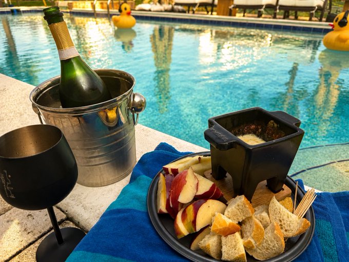 A bottle of champagne chills in an ice bucket beside a wine glass and a snack board of bread, apple slices, and dip, all set poolside next to two yellow rubber ducks floating in the water—perfectly capturing the simple luxury of savoring life's small pleasures.