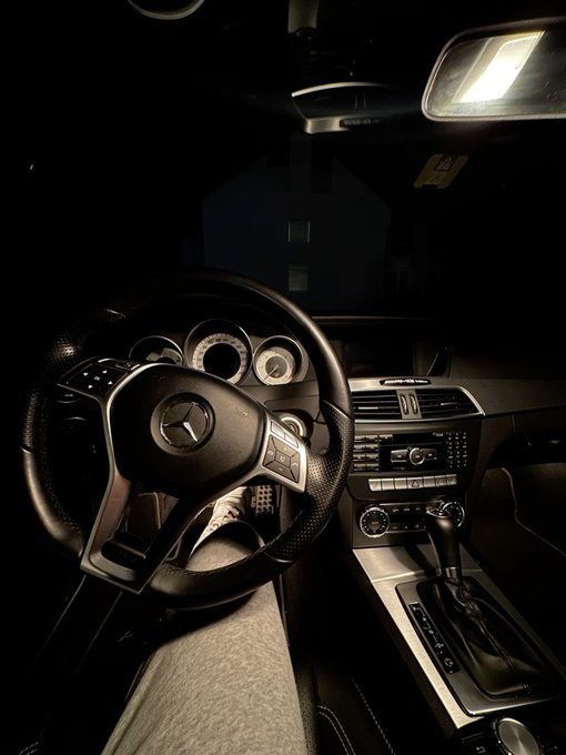 Interior of a Mercedes-Benz vehicle at night, showing the steering wheel with the Mercedes logo, dashboard gauges, center console with gear shifter, and a dimly lit house visible through the windshield.