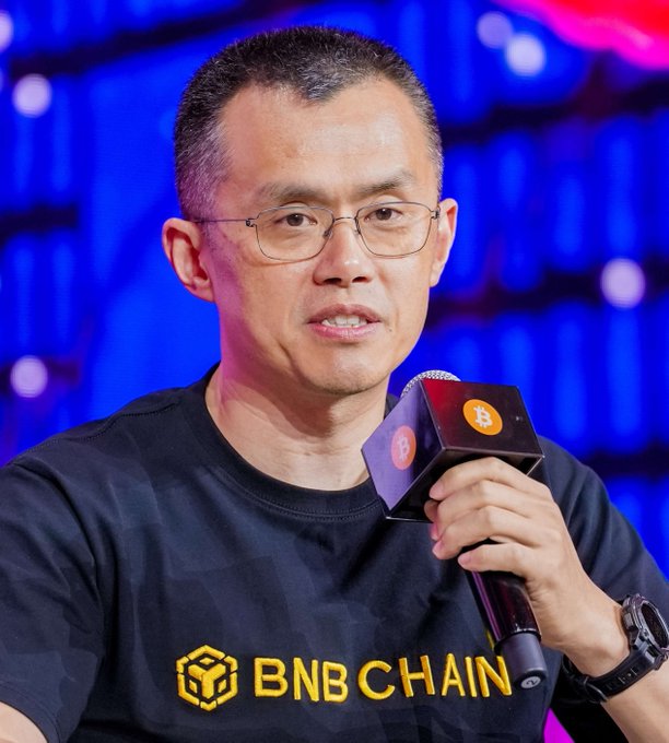 A man wearing a "BNB CHAIN" shirt speaks into a microphone branded with the Bitcoin logo, visually aligning with CZ's playful post about waiting for sensationalist Bitcoin crash headlines as the price dips from $1,001,000 to $985,000.