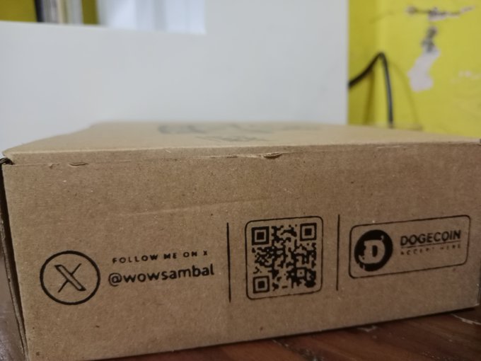 This image shows a cardboard box with a QR code and a Dogecoin logo, indicating that WOW Sambal accepts Dogecoin for payment and encourages customers to support the cryptocurrency.