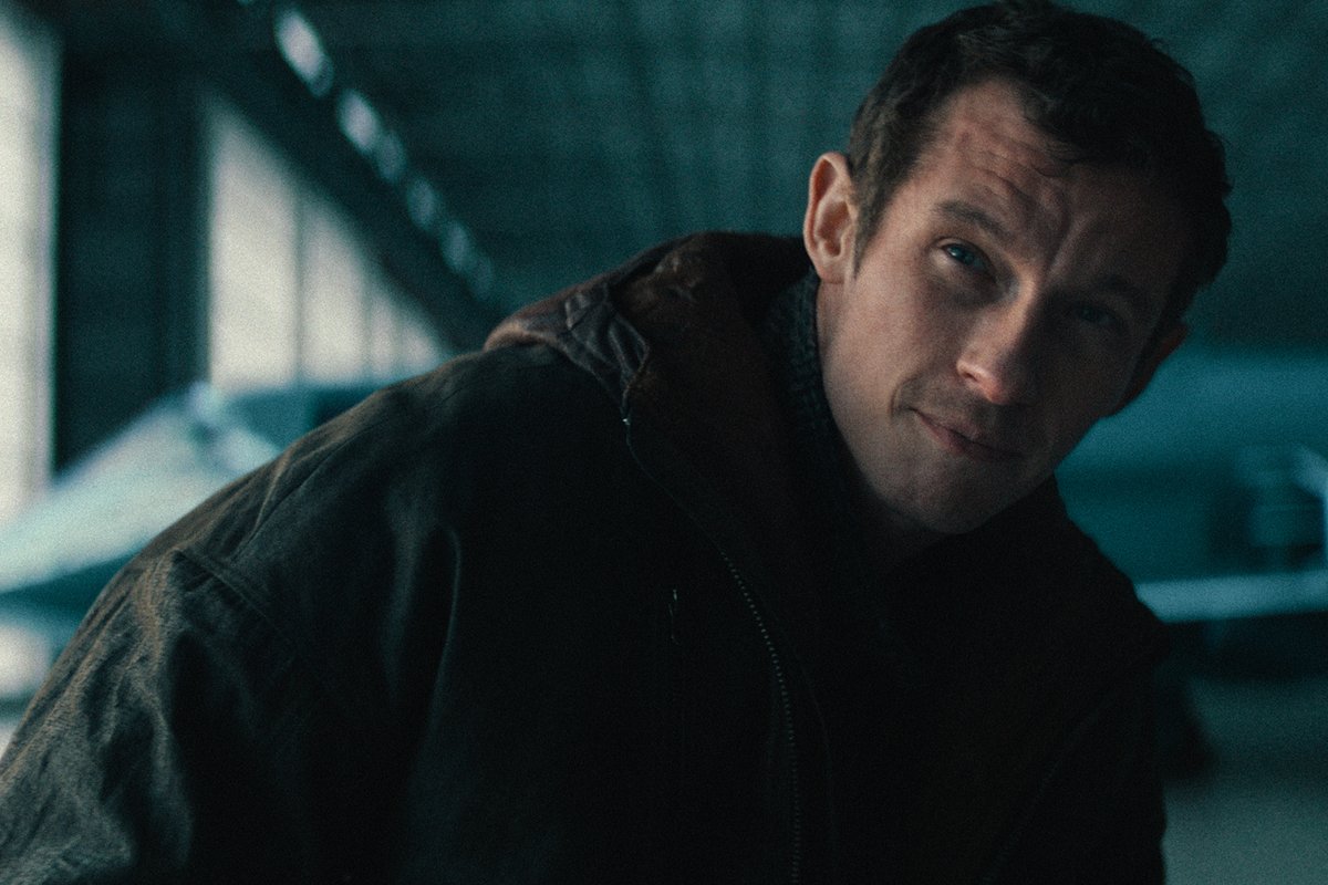 A cinematic close-up shot of actor Callum Turner, who plays Newt in the 'Fantastic Beasts' and 'Harry Potter' films, wearing a dark jacket against a blurred blue background.