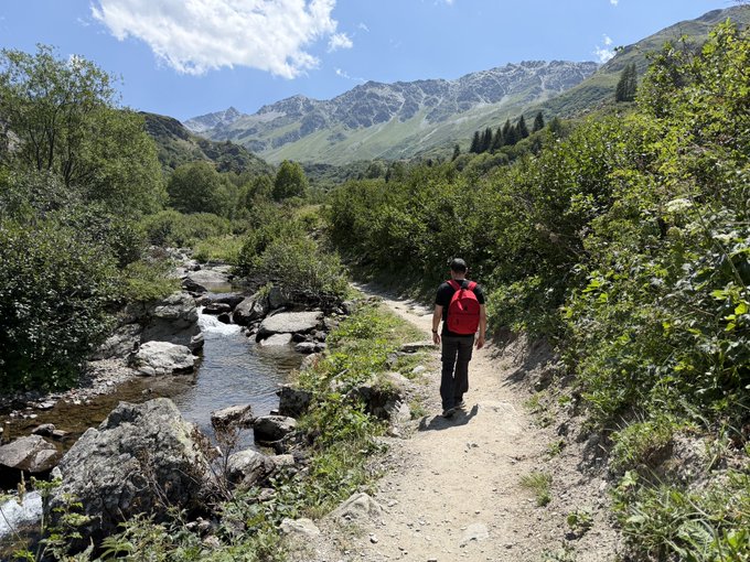 A scenic mountain landscape with a hiker wearing a red backpack walking along a dirt trail beside a rocky stream, surrounded by green vegetation and snow-dusted peaks under a partly cloudy sky.