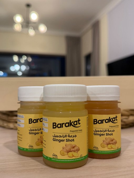 Three bottles of "Barakat" brand "Ginger Shot" prepared daily drink are arranged on a table in a softly lit room.