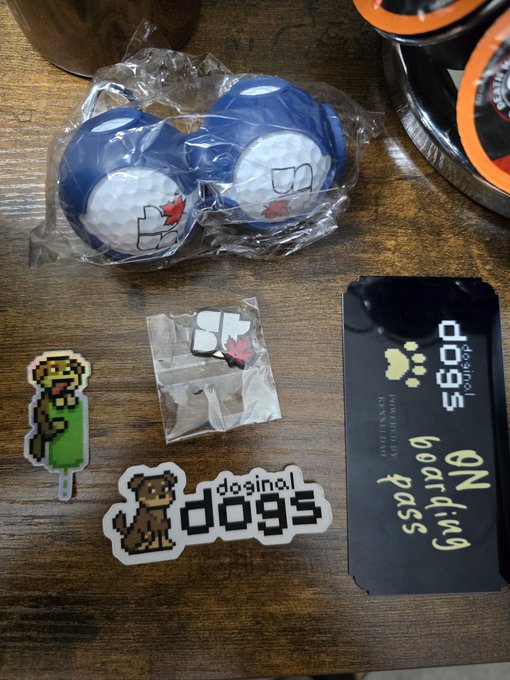 A collection of merchandise and promotional items on a wooden table, including blue and white golf balls in plastic packaging, pixel-art stickers, and a black card reading 'doginal dogs ON board ding pass'.