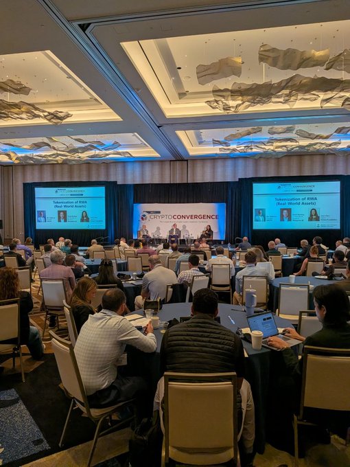 The image shows a large conference room filled with people sitting at tables, engaged in a panel discussion or presentation. The attendees are seated on chairs arranged around the tables, which have laptops open on them. Some of the individuals appear to be taking notes during the event. A few people can also be seen holding cups and handbags.

The room is well-lit with multiple large screens mounted on the walls, likely used for presentations or visual aids. The overall atmosphere suggests a professional gathering focused on discussing topics such as crypto, tokenization, digital assets, and portfolio construction in the context of the rapidly evolving financial landscape.