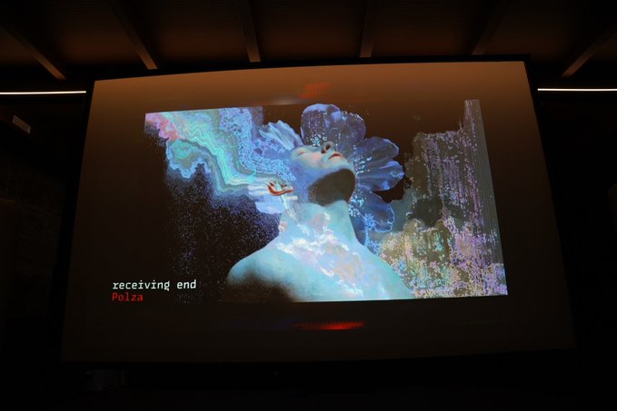 A video art piece titled "receiving end" by Polza, displayed on a large projection screen in a dimly lit gallery space, featuring a surreal figure with a crystalline, flower-like headdress against an iridescent backdrop—presented as part of the artist's Venice Biennale 2026 exhibition available on @objktcom.