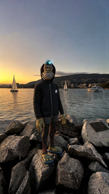 A costumed figure wearing a shark-like mask with a headlamp and yellow gloves stands atop rocky shoreline at sunset, with sailboats and a yacht visible on the water behind. The image pairs with the "GM to those that sail through the storm" message by symbolizing resilience—standing firm on the rocks while vessels navigate the tranquil yet challenging waters of dawn or dusk.