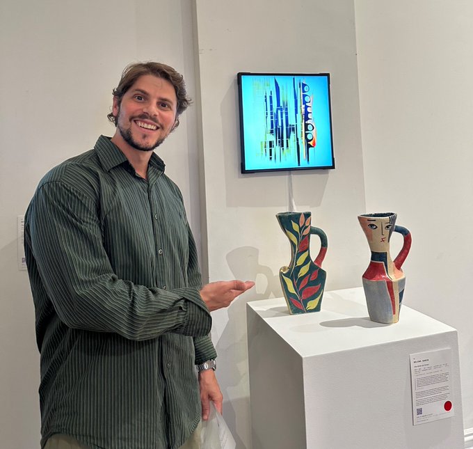 A smiling man in a green striped shirt standing in a gallery next to two colorful ceramic pitcher sculptures on a white pedestal, with a digital artwork displayed on the wall behind them.