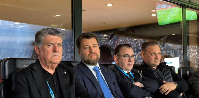 Four men sit in stadium VIP seats watching a football match, with one wearing a lanyard and another in a suit and tie, while a screen displays match footage in the background. The Turkish post humorously suggests pressing the TLY (Turkish Lira) button instead of asking Emre for money, seemingly poking fun at a person named Emre in the image.