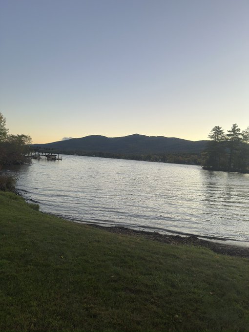A serene lakeside scene at dusk features calm water reflecting the soft sky, with silhouetted mountains in the background and a dock on the left shore.