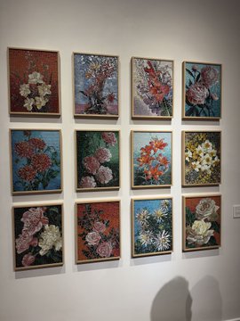 A gallery wall displaying a grid of twelve framed floral paintings featuring various flowers like roses, hollyhocks, and lilies against muted backgrounds.