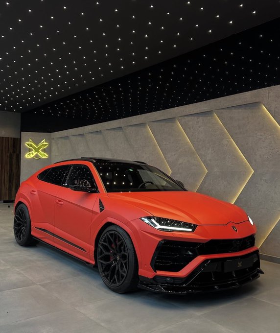 A matte red Lamborghini Urus SUV parked in a showroom with a starry ceiling, black wheels, and aggressive body styling.