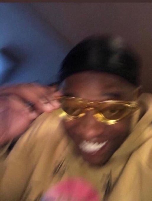 A blurry close-up of a smiling man wearing yellow-tinted sunglasses and a tan hoodie, with his hand near his face in a celebratory gesture. The joyful, triumphant expression perfectly captures the excitement of unexpectedly profiting from a risky, uninformed investment.