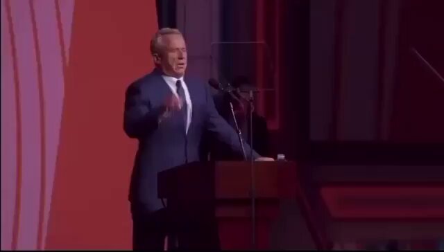 The image features a man standing at a podium with his hands raised while speaking into a microphone. The background of the image has red and purple stripes, which could be an artistic choice or part of the stage setup for an event. The caption "RFK Jr." is written in white text above the speaker's head, indicating that he was delivering a speech at a political event.