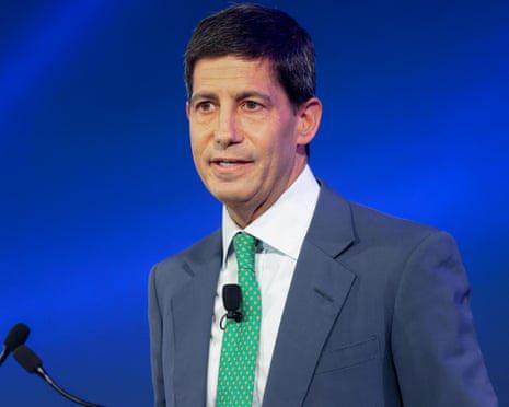 A man in a blue suit with a green tie speaking at a podium against a blue background.
