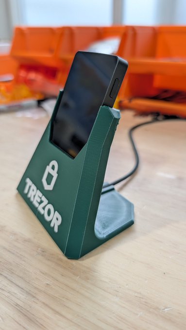 The image features a green Trezor phone holder that has been designed by someone else. The phone holder is placed on top of a wooden table, with an orange box behind it. There are two other orange boxes visible in the background as well. A caption below the image reads "As long as you opensource the design and share it with everyone, you are fine. Right @Trezor ?". This message seems to be related to the Trezor phone holder that is placed on a wooden table.