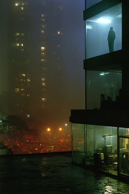 A moody, cinematic nighttime scene showing a silhouetted figure standing in a brightly lit glass office on the upper floor of a building, with a foggy cityscape and illuminated skyscrapers in the background.
