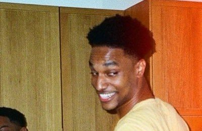 A young Black man smiling while glancing back at the camera in what appears to be a casual indoor setting with wooden cabinets in the background.