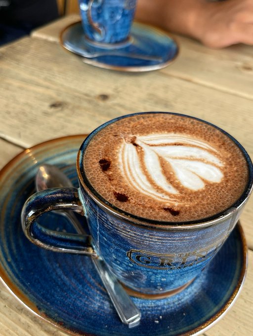 A blue ceramic coffee cup labeled "CRY" with latte art sits on a matching saucer on a wooden table, with a second cup blurred in the background.