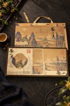 Two traditional Chinese scroll-style artworks or folders depicting landscape paintings and calligraphy, displayed on a dark surface with tea and plants nearby.