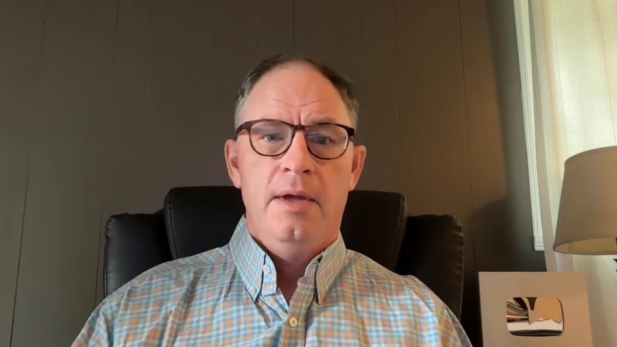 The image shows a man wearing glasses sitting in an office chair, looking at the camera with a serious expression. Behind him, there is a lamp that provides lighting for his workspace. The background of the image features a gray wall, which contrasts with the white frame around the picture. In the bottom right corner of the image, there is a link to a YouTube video titled "Ripple Is The One Stop Crypto Infrastructure Stop Watch The Full Youtube Video Here: youtu.be/Dt9uE-KHGao". This suggests that the man might be discussing or reviewing the Ripple cryptocurrency platform in his office setting.