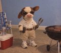 A small mascot character wearing sunglasses, a tan jacket, and a backpack stands next to a grill and a red cooler.