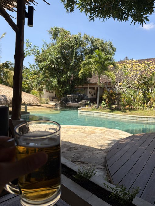 A hand holds a glass mug of beer in the foreground, overlooking a tranquil swimming pool surrounded by tropical trees and a villa under a clear blue sky.