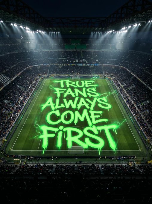 The image shows a large stadium with green graffiti that says "True fans always come first" written on the field. The text is in white, making it stand out against the green background of the soccer pitch. The stadium appears to be empty at night, adding to the overall atmosphere and emphasizing the message of unity and dedication among the fans.
