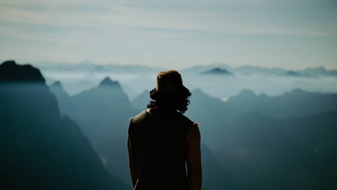 A person wearing a hat viewed from behind, standing on a mountain overlook and gazing out at a vast range of misty, blue-tinted peaks.