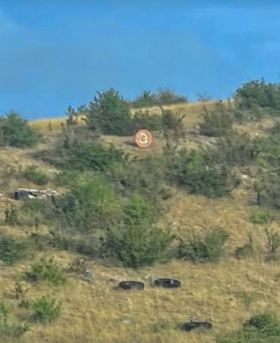 A hillside view featuring a large orange Bitcoin "₿" symbol among scattered shrubs and vintage tires, visually representing the "Spot the #Bitcoin" caption in a rural French landscape.