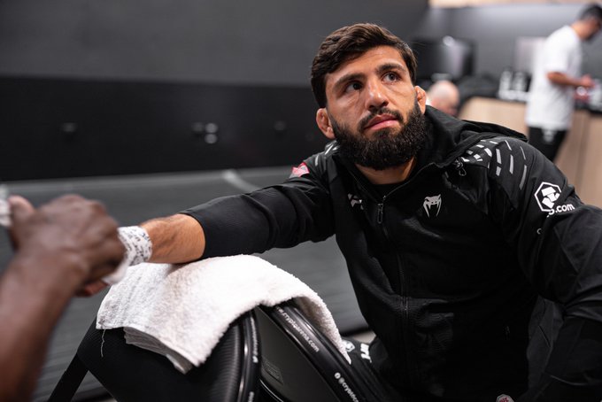 A bearded fighter in a black Venum hoodie sits with a towel draped over his knee, reaching out to fist bump someone while looking up in concentration. The "Crypto.com" logo on his sleeve matches the #CryptoCom sponsor branding referenced in the post about the upcoming Arman Tsarukyan vs. Mauricio Ruffy bout.