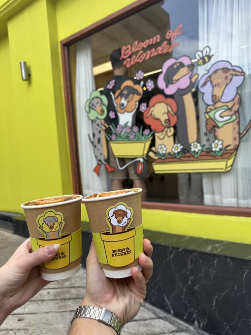 Two hands holding paper cups labeled "BOBBY & FRIENDS" with cartoon dog illustrations, held up in front of a storefront window featuring "Bloom of Wonder" artwork with various dogs and flowers.