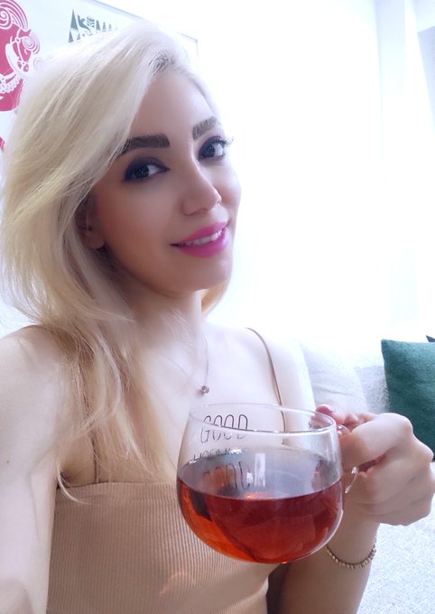 A blonde woman with pink lipstick smiles while holding a glass mug filled with a reddish beverage, wearing a beige tank top.