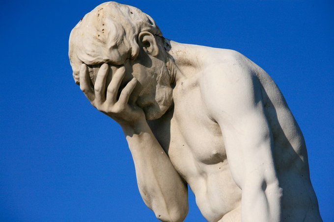 A white marble statue depicts a nude muscular man covering his face with his hand in a gesture of despair or contemplation, set against a clear blue sky.