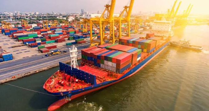 An aerial view captures a massive cargo ship loaded with colorful shipping containers docked at a busy port during golden hour.
