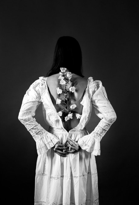 A black-and-white artistic photograph shows a woman from behind, wearing a flowing white dress with an open back decorated with delicate flowers climbing up her spine. The ethereal, bridal aesthetic of the image visually represents the "Paper Brides" NFT collection mentioned in the post, where the artist expresses gratitude to collectors supporting their project.