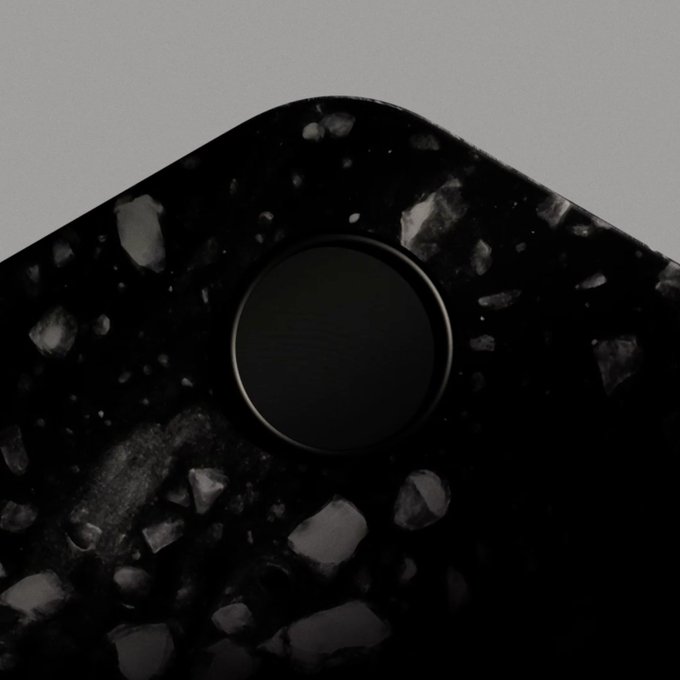 A close-up of a sleek black speckled phone case or hardware surface featuring a circular camera module, its dark, rugged aesthetic reinforcing the "Hardware built for hard money" tagline by evoking a sense of durable, premium quality.